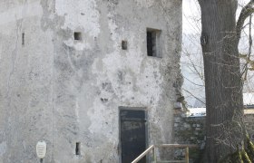 Ein alter Turm mit moosbedecktem Dach, daneben ein Baum. Drei Personen sitzen an einem Tisch, w&auml;hrend zwei Personen vorbeigehen.