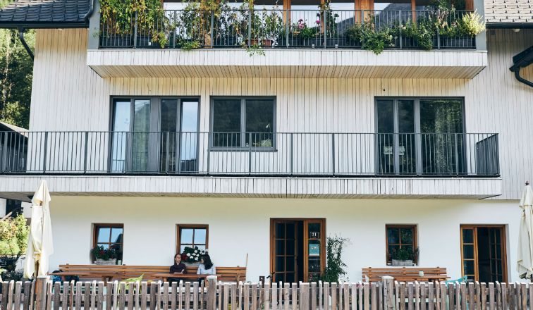 Ein modernes dreist&ouml;ckiges Haus mit Holzbalkonen und Pflanzen. Vor dem Haus sitzen zwei Personen auf einer Bank hinter einem Holzzaun.