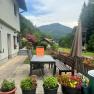 Terrasse einer Ferienwohnung mit Tisch, St&uuml;hlen und Blumen, umgeben von gr&uuml;nen H&uuml;geln und Wald.