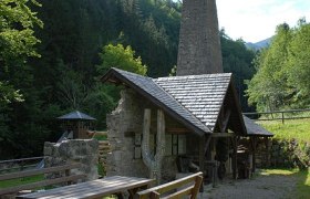 Historische Schmiede mit Kamin im Wald, Holztische im Vordergrund.
