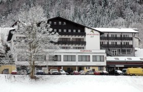Ein verschneites Hotelgeb&auml;ude mit der Aufschrift 'Sporthotel' und 'Cafeteria', umgeben von schneebedeckten B&auml;umen und geparkten Autos.