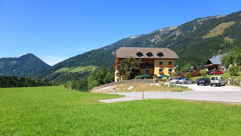 Ein gelbes Gebäude mit Balkon und Blumen, umgeben von grünen Wiesen und Bergen unter blauem Himmel.