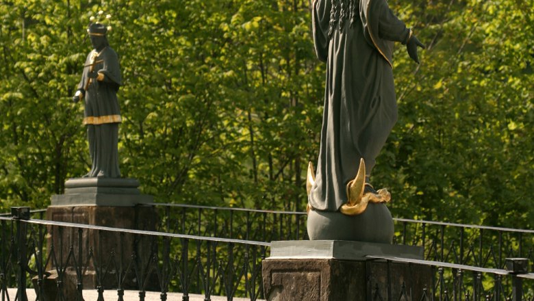 Zwei Statuen auf einer Br&uuml;cke mit Gel&auml;nder, umgeben von gr&uuml;nen B&auml;umen.