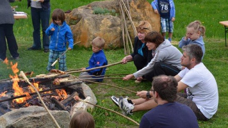 Menschen sitzen um ein Lagerfeuer und rösten Brot an langen Stöcken.
