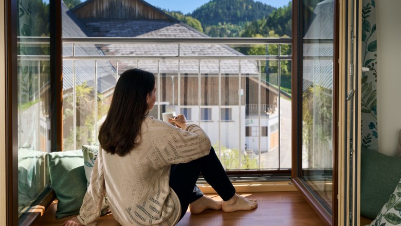Eine Frau sitzt auf einer Fensterbank und blickt auf eine l&auml;ndliche Landschaft mit Bergen im Hintergrund.