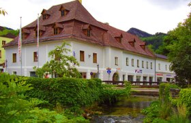 Historisches Geb&auml;ude mit rotem Dach an einem Bach, umgeben von gr&uuml;ner Vegetation.