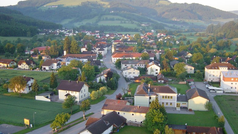Luftaufnahme eines Dorfes mit Häusern, Straßen und umliegenden Hügeln im Hintergrund.