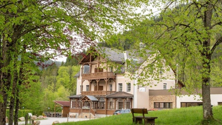 Fastenhaus Maria Seesal, © Fastenhaus Ein großes Gebäude mit Holzbalkonen, umgeben von grünen Bäumen und einer Wiese mit einer Bank im Vordergrund.