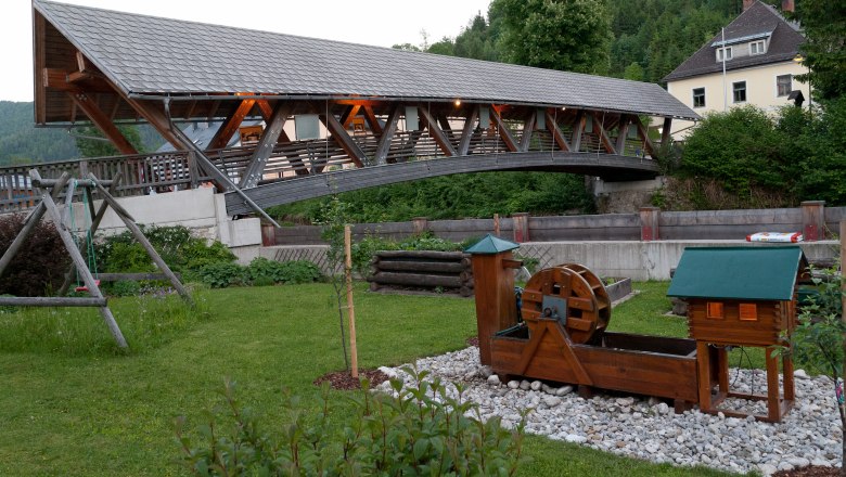 &Uuml;berdachte Holzbr&uuml;cke &uuml;ber einen Fluss mit einem kleinen Wasserrad im Vordergrund.