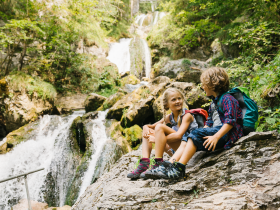 Zwei Kinder sitzen entspannt auf einem gro&szlig;en Stein und genie&szlig;en die erfrischende Atmosph&auml;re des Naturparks. Umgeben von &uuml;ppigem Gr&uuml;n und dem sanften Rauschen des Wassers, strahlt die Szene pure Lebensfreude und Abenteuerlust aus.