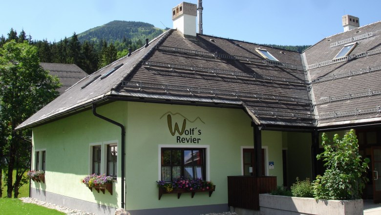 Ein gr&uuml;nes Geb&auml;ude mit der Aufschrift 'Wolf's Revier' und Blumenk&auml;sten vor den Fenstern, umgeben von B&auml;umen und Bergen im Hintergrund.