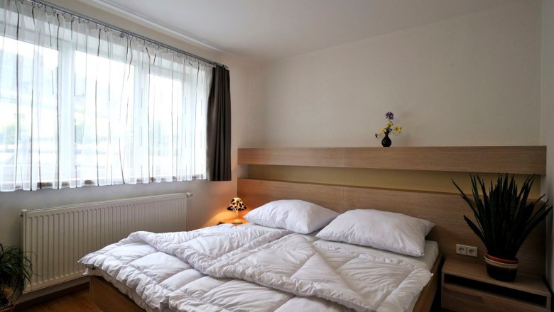 Modern eingerichtetes Schlafzimmer mit Doppelbett, hellem Holzmobiliar und großen Fenstern mit weißen Vorhängen.