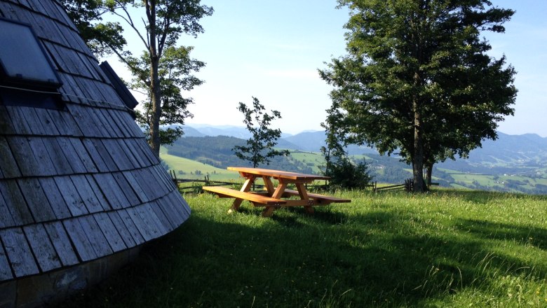 Holzh&uuml;tte und Picknicktisch auf einer Wiese mit B&auml;umen und Bergblick.