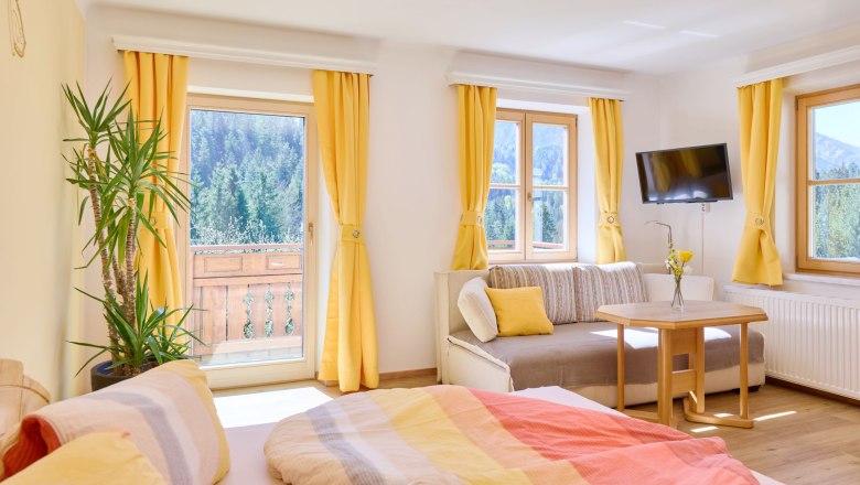 Heller Raum mit Bett, Sofa, Tisch und gelben Vorh&auml;ngen, Blick auf Wald und Berge.